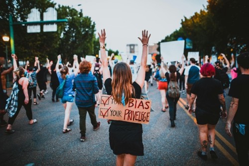 Protesters walking away from camera. On persons back is the sign that says check your bias, check your privilege