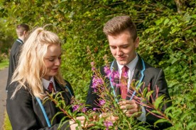 two young people wearing blazers stand before a spray of pink flowers with a backdrop of hedgerow. They smile and look down at the flowers