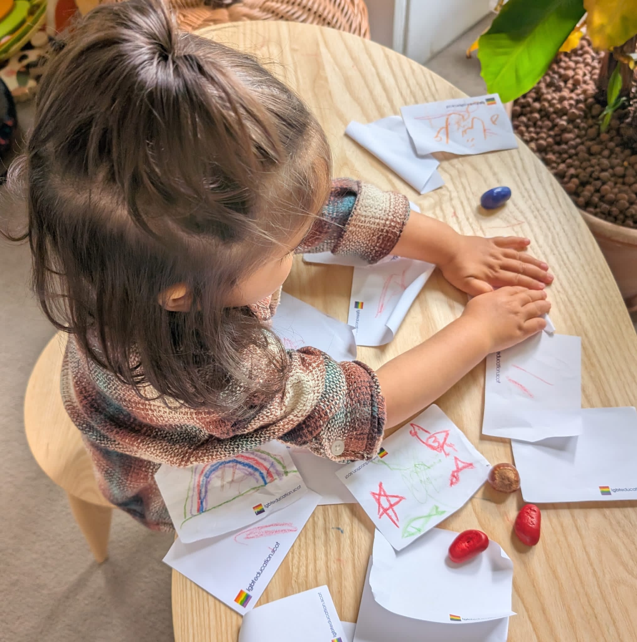 Young child sits at a table with several crayon drawings of stars and a rainbow on LGBT education notepaper